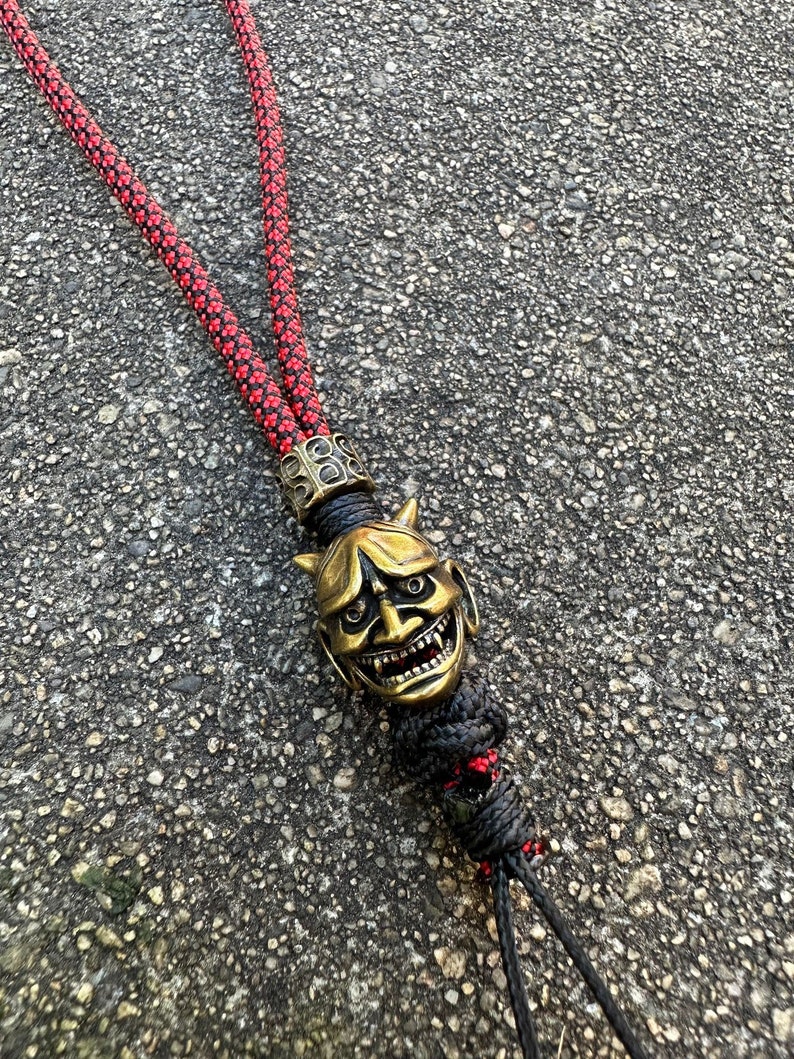 May include: A red and black paracord lanyard with a gold-coloured oni mask charm. The mask has a fierce expression with horns, sharp teeth, and red eyes.