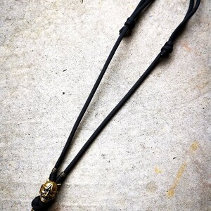 May include: A black paracord necklace with a gold-colored skull pendant. The skull has a detailed design with horns and a menacing expression.