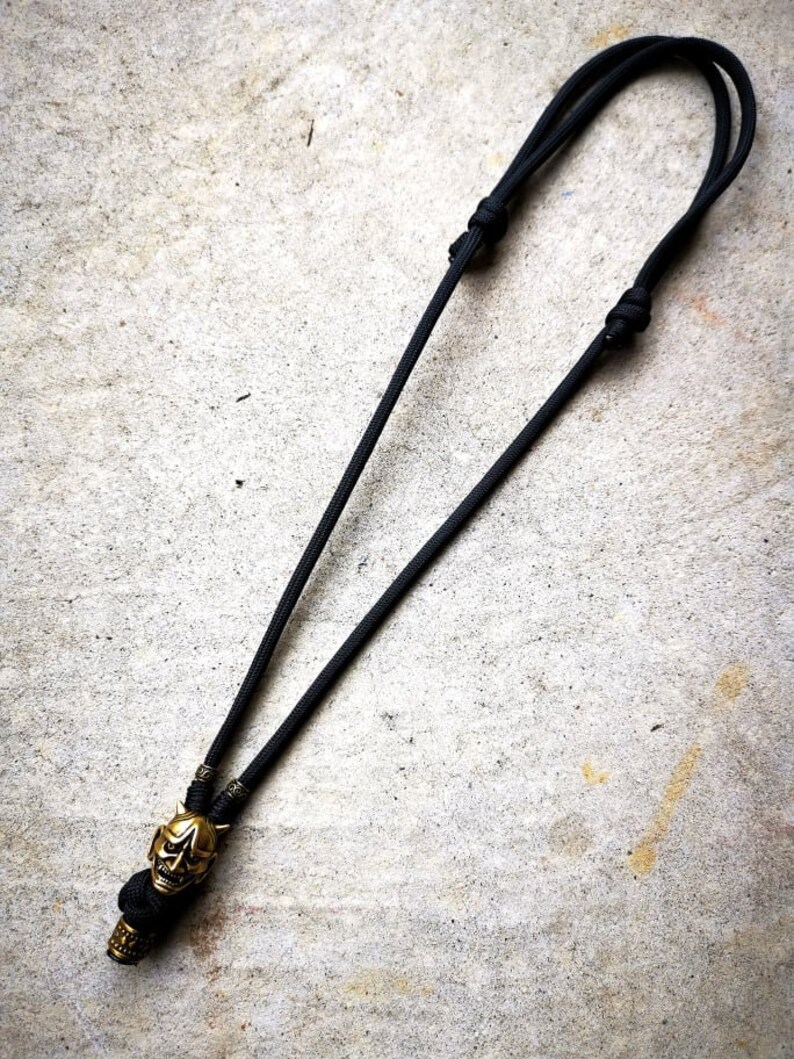 May include: A black paracord necklace with a gold-colored skull pendant. The skull has a detailed design with horns and a menacing expression.