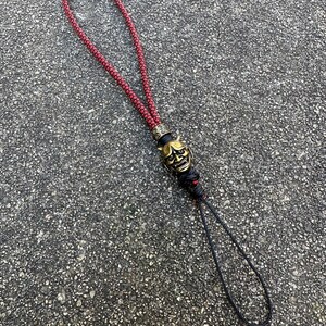 May include: A red and black braided lanyard with a gold-coloured oni mask charm. The lanyard is made of a thick, durable cord and has a loop at the top for attaching to a key chain or other item.