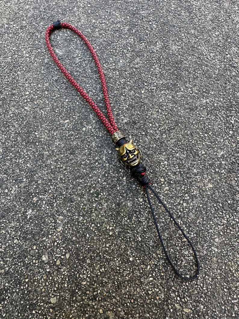 May include: A red and black braided lanyard with a gold-coloured oni mask charm. The lanyard is made of a thick, durable cord and has a loop at the top for attaching to a key chain or other item.