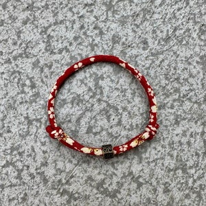 Puede incluir: Pulsera roja y blanca con estampado floral y una peque&ntilde;a cuenta negra cuadrada. La pulsera est&aacute; hecha de un material tejido y est&aacute; anudada en los extremos. La pulsera tiene forma circular.