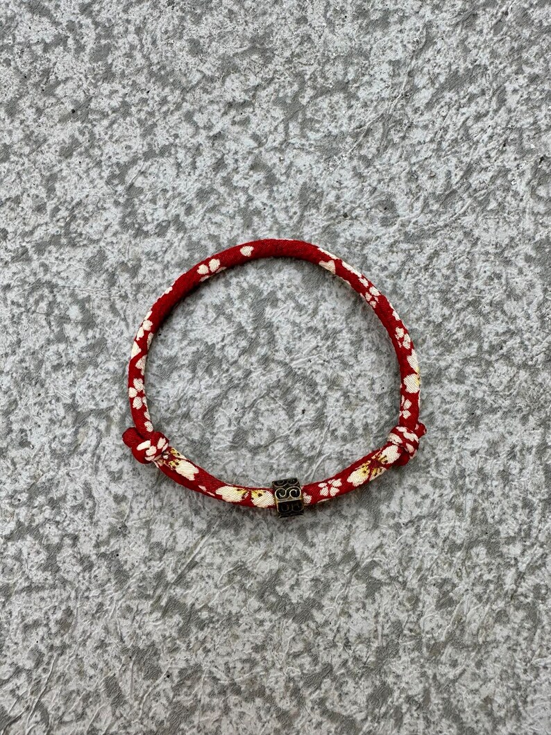 Puede incluir: Pulsera roja y blanca con estampado floral y una peque&ntilde;a cuenta negra cuadrada. La pulsera est&aacute; hecha de un material tejido y est&aacute; anudada en los extremos. La pulsera tiene forma circular.