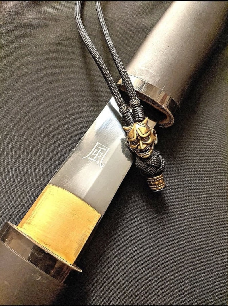 May include: A black sword with a gold-colored hilt and a black cord with a gold-colored oni mask charm. The sword has the kanji for "wind" inscribed on the blade.