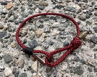 May include: A red and black rope bracelet with a silver clasp and a brass charm. The bracelet is lying on a bed of small gray stones.