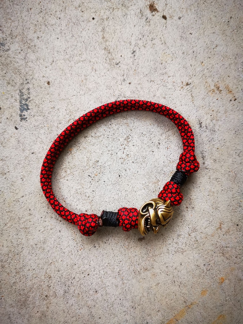 Op de afbeelding: Een rood en zwart paracord armband met een gouden metalen bedel in de vorm van een wolfskop.