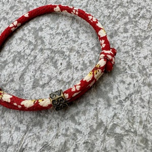 Puede incluir: Una pulsera con estampado floral rojo y blanco con un acento met&aacute;lico de color bronce. La pulsera est&aacute; hecha de un material tejido y presenta un dise&ntilde;o floral repetitivo. El acento met&aacute;lico es una peque&ntilde;a cuenta cuadrada con un patr&oacute;n intrincado.