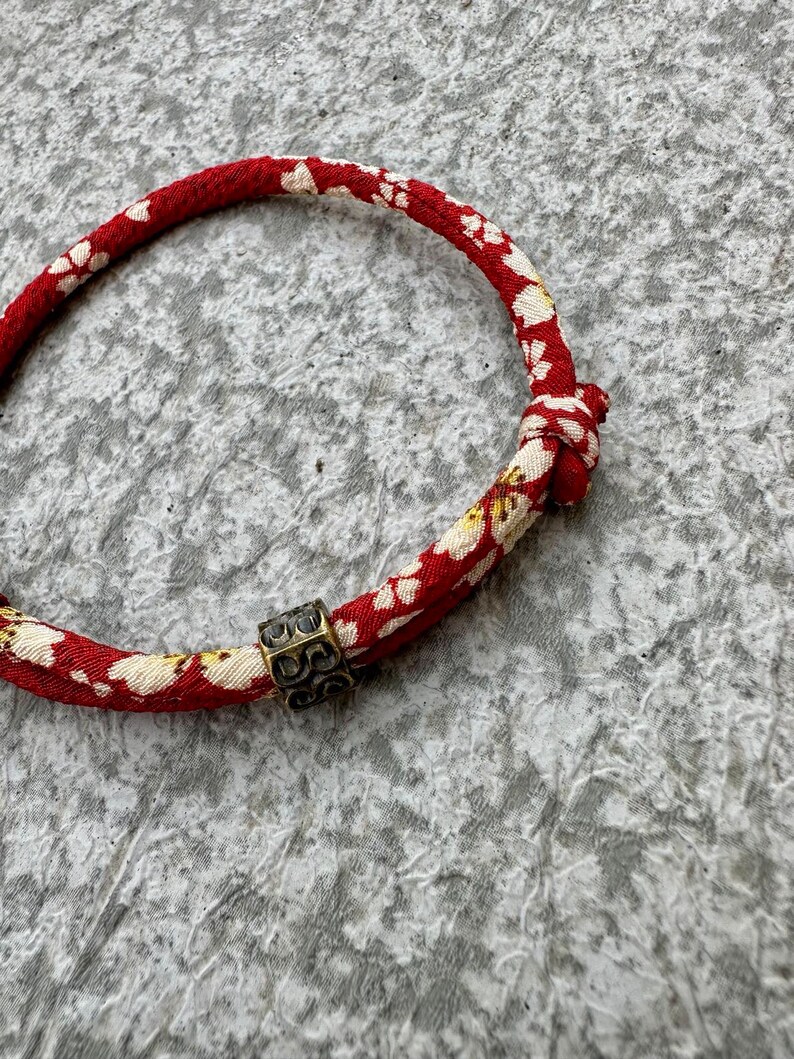 Puede incluir: Una pulsera con estampado floral rojo y blanco con un acento met&aacute;lico de color bronce. La pulsera est&aacute; hecha de un material tejido y presenta un dise&ntilde;o floral repetitivo. El acento met&aacute;lico es una peque&ntilde;a cuenta cuadrada con un patr&oacute;n intrincado.