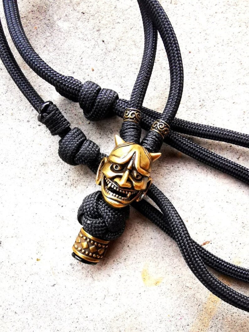 May include: A black paracord necklace with a gold-colored oni mask pendant and a decorative bead. The pendant features a detailed design of a demon face with horns and fangs.