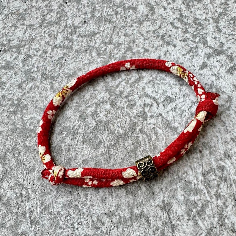 Puede incluir: Pulsera roja y blanca con estampado floral y un acento met&aacute;lico dorado. La pulsera est&aacute; hecha de un cord&oacute;n cubierto de tela y est&aacute; atada en los extremos. La pulsera tiene aproximadamente 18 cm de circunferencia.