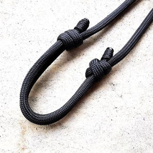 May include: A black paracord lanyard with two knots tied in it. The lanyard is on a white surface.