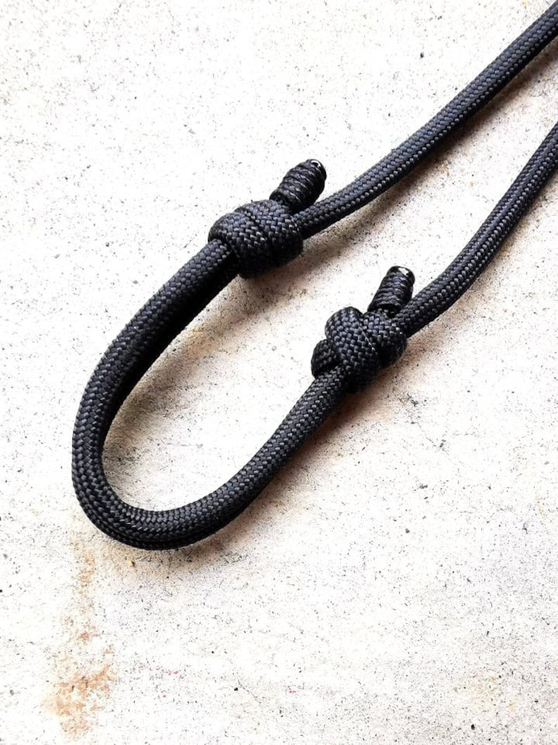 May include: A black paracord lanyard with two knots tied in it. The lanyard is on a white surface.