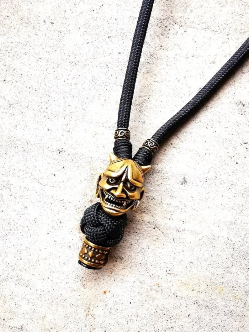 May include: A black paracord necklace with a gold-colored oni mask bead and two decorative metal beads.
