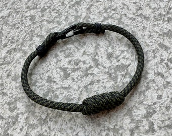 May include: A green and black paracord bracelet with a black metal clasp. The bracelet is tied with a knot on one end.