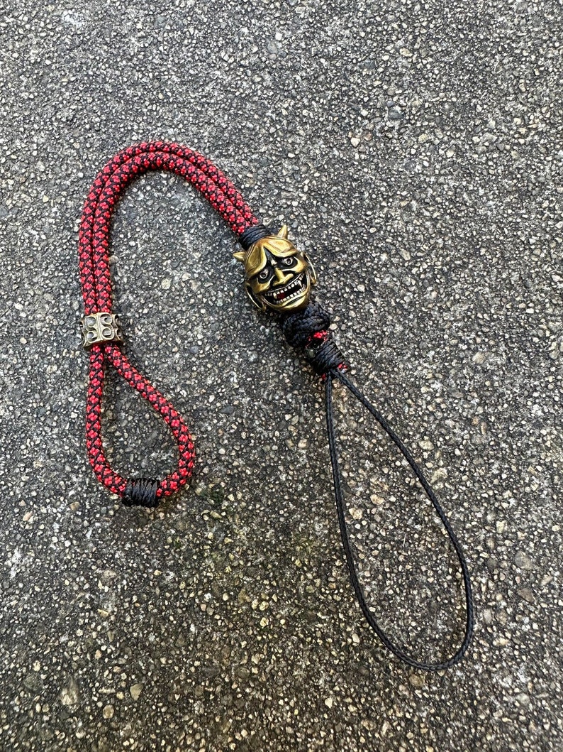May include: A red and black paracord lanyard with a gold-coloured oni mask charm. The lanyard has a loop at the top and a knot at the bottom.