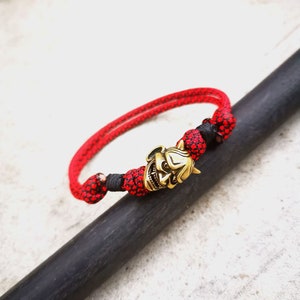 Op de afbeelding: Een rood en zwart gevlochten armband met een gouden oni-masker bedel.