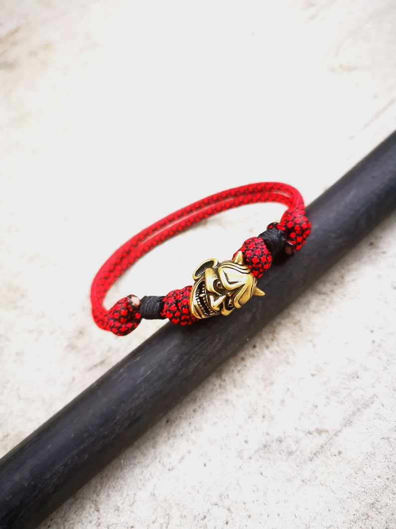 Op de afbeelding: Een rood en zwart gevlochten armband met een gouden oni-masker bedel.