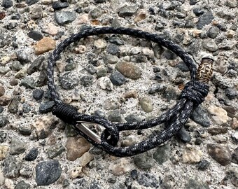 May include: A black and gray rope bracelet with a silver clasp and a decorative bronze bead. The bracelet is lying on a textured, gray and brown stone surface.