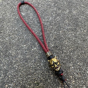 May include: A red and black rope lanyard with a gold-coloured oni mask charm. The mask has a fierce expression with horns and sharp teeth.
