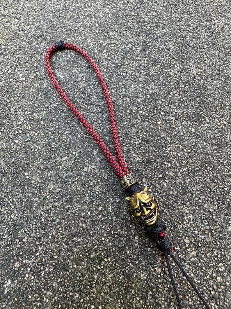 May include: A red and black rope lanyard with a gold-coloured oni mask charm. The mask has a fierce expression with horns and sharp teeth.
