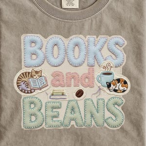 以下が含まれることがあります： 「BOOKS and BEANS」と書かれたグラフィックが特徴のグレーのComfort Colors Tシャツ。グラフィックには、本を読む猫、コーヒーカップ、コーヒー豆、本が含まれています。文字は水色、ピンク、緑色です。