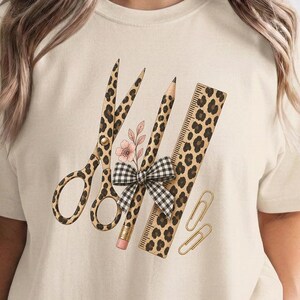May include: A cream-coloured t-shirt featuring a graphic print of scissors, a pencil, and a ruler, all decorated with a brown and black leopard print pattern. A small pink flower and a black and white gingham bow are centred on the pencil.