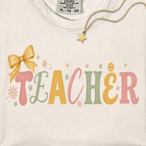 May include: A cream-colored t-shirt with a Comfort Colors label at the neck. The front features the word TEACHER in large, multi-colored letters with a yellow bow, small flowers, stars, and a smiley face icon integrated into the design.