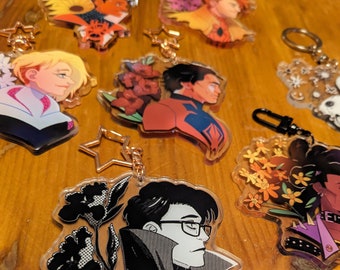 May include: A set of seven black and white anime-style character keychains with floral accents. Each keychain features a different character with a unique design and a star-shaped charm.