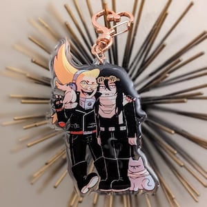May include: A clear acrylic keychain featuring a cartoon illustration of two characters in black outfits. The keychain has a rose gold heart-shaped clasp. One character has blonde hair and the other has black hair. A white cat is also featured.