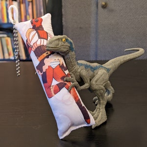 Puede incluir: Una almohada blanca con una ilustración colorida de un personaje de superhéroe. Un juguete de dinosaurio azul y gris muerde la almohada.