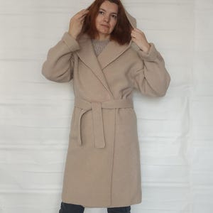 Puede incluir: Un abrigo beige con capucha, mangas largas con puños vueltos, solapa ancha y un cinturón de lazo a juego en la cintura. El abrigo se lleva sobre un suéter gris texturizado, pantalones gris oscuro y zapatos sin cordones gris oscuro con suela blanca.