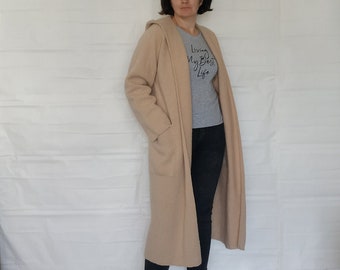 May include: A long, beige cardigan with a hood and pockets. The cardigan is open and the person wearing it is standing with their hands in the pockets. The person is wearing a grey t-shirt with the text "Living My Best Life" printed on it. The person is also wearing dark blue jeans and grey sneakers.