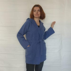 Könnte beinhalten: Ein blauer Kapuzenmantel mit passendem Gürtel und Taschen. Der Mantel ist knielang und aus weichem Material. Die Person trägt eine dunkelgraue Hose und schwarze Slip-on-Schuhe.