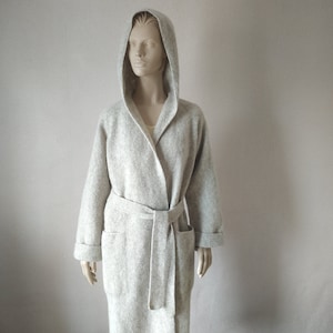 Puede incluir: Una bata con capucha de color gris claro con cinturón y bolsillos a juego. La bata está hecha de un material suave y texturizado y tiene mangas largas con puños doblados. El maniquí lleva una camiseta de color claro debajo.