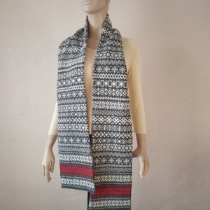 May include: A gray and white patterned scarf with a red stripe. The scarf is made of a soft, knitted fabric.