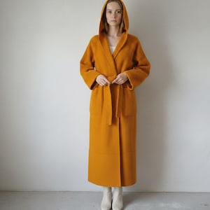 Könnte beinhalten: Langer, kapuzenartiger Robe in Senffarbe mit passendem Gürtel. Die Robe hat lange Ärmel und Taschen. Die Person trägt weiße Stiefel. Die Robe ist aus einem weichen, strukturierten Material.