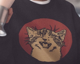 May include: A black sweatshirt with a red circle and a cartoon image of a cat with its mouth open, as if laughing. The cat is in a light brown color.