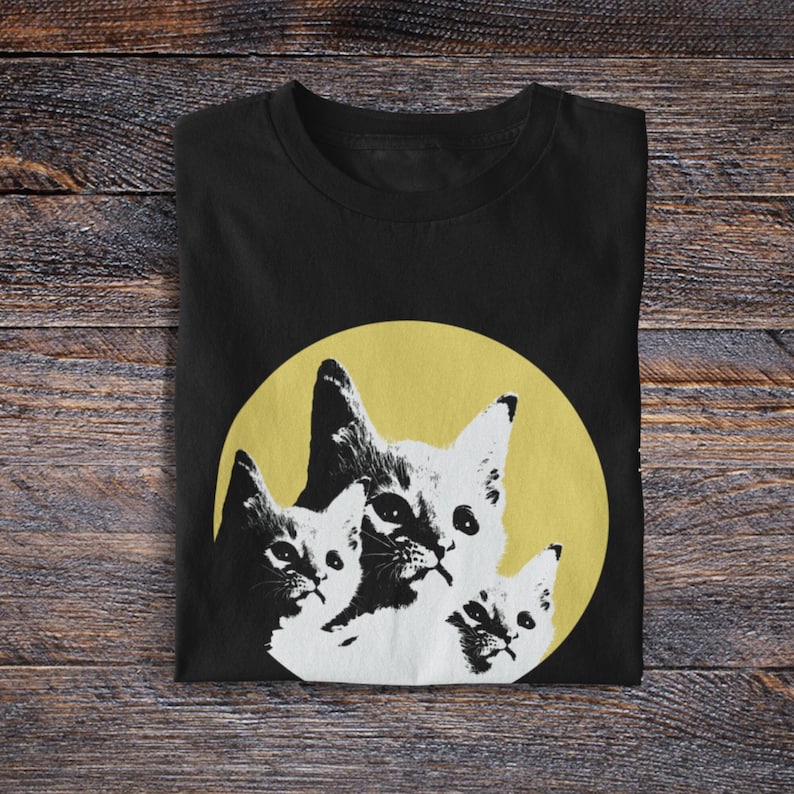 Pode incluir: Camiseta preta com um gr&aacute;fico de tr&ecirc;s gatos em um c&iacute;rculo amarelo. Os gatos est&atilde;o empilhados um em cima do outro, com o gato maior na parte inferior e o gato menor na parte superior.