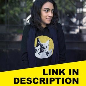 Pode incluir: Uma mulher vestindo uma camisa preta de manga comprida com um gr&aacute;fico de tr&ecirc;s gatos em um c&iacute;rculo amarelo. A camisa tem uma echarpe preta ao redor do pesco&ccedil;o.