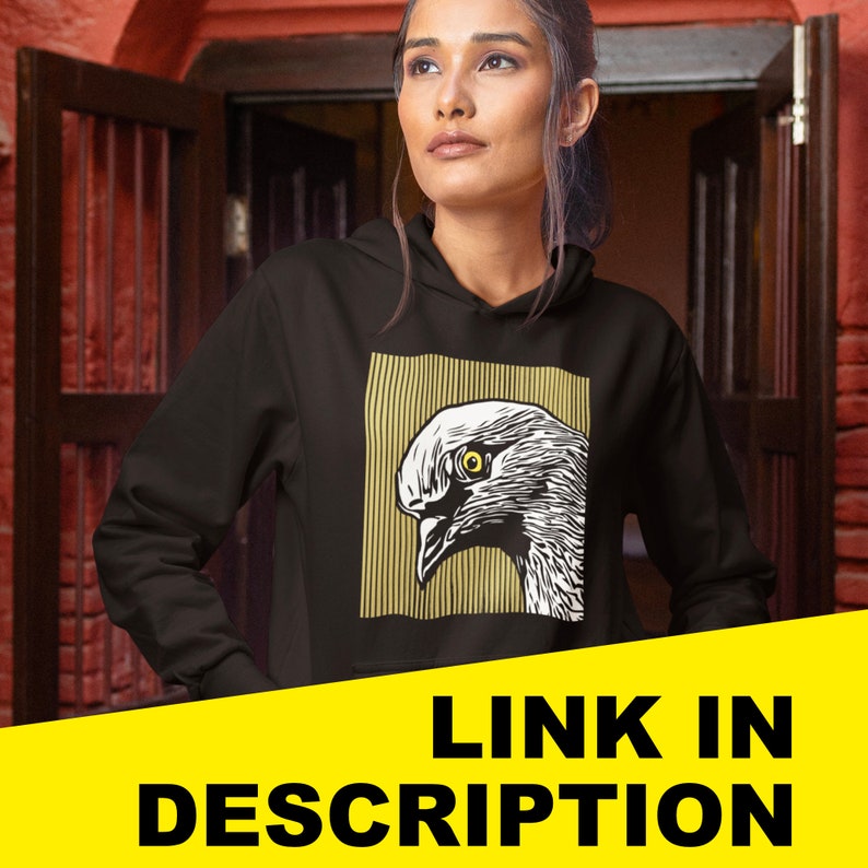 May include: Black hoodie featuring a graphic print of a white bird with a yellow eye. The bird is enclosed within a yellow and white striped border. The text "LINK IN DESCRIPTION" is printed in yellow on a black background.