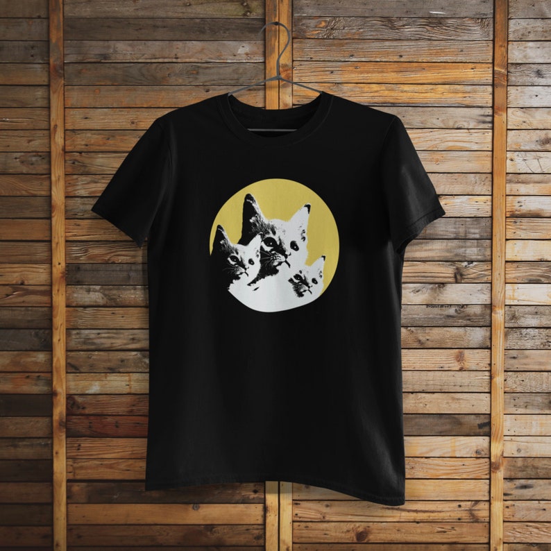Pode incluir: Camiseta preta com um gr&aacute;fico de tr&ecirc;s rostos de gatos em um c&iacute;rculo amarelo. Os gatos est&atilde;o olhando para fora do c&iacute;rculo.