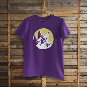 Pode incluir: Camiseta roxa com um gr&aacute;fico de tr&ecirc;s rostos de gatos em um c&iacute;rculo amarelo.