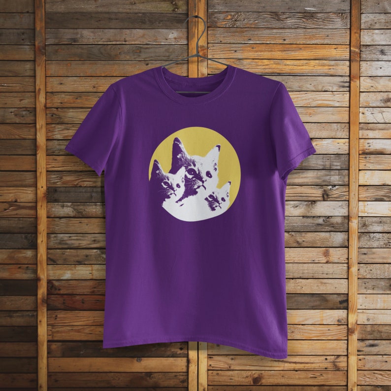 Pode incluir: Camiseta roxa com um gr&aacute;fico de tr&ecirc;s rostos de gatos em um c&iacute;rculo amarelo.