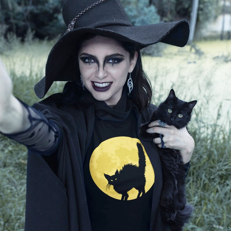 May include: A woman wearing a black witch hat and a black shirt with a yellow moon and black cat silhouette. She is holding a black cat in her arms.