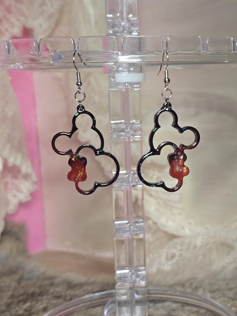May include: A pair of dangle earrings with a dark grey outline in a Mickey Mouse shape. Each earring has a small, reddish-orange charm inside the outline. The earrings hang from silver-coloured fish hook ear wires.