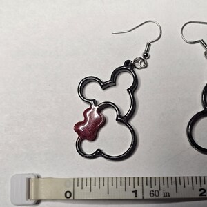 May include: A pair of dark grey Mickey Mouse-shaped earrings with a red, glittery accent. The earrings are shown next to a measuring tape, with the earrings measuring approximately 2.5 cm in length.