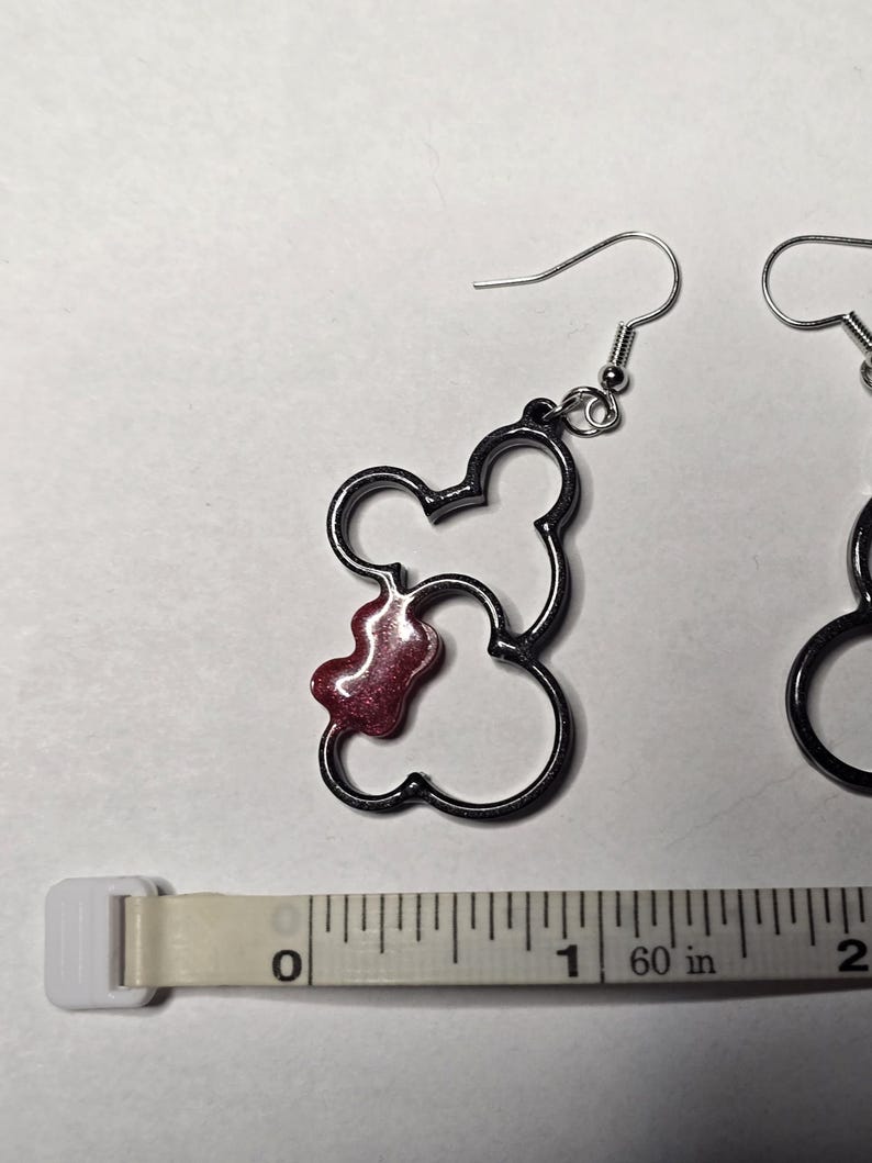 May include: A pair of dark grey Mickey Mouse-shaped earrings with a red, glittery accent. The earrings are shown next to a measuring tape, with the earrings measuring approximately 2.5 cm in length.