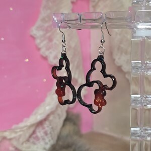 May include: A pair of dark brown, Mickey Mouse-shaped dangle earrings with a glittery outline, suspended from silver-coloured fish hook ear wires. The background is a soft pink. The earrings are a fun accessory.
