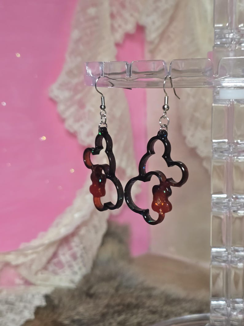 May include: A pair of dark brown, Mickey Mouse-shaped dangle earrings with a glittery outline, suspended from silver-coloured fish hook ear wires. The background is a soft pink. The earrings are a fun accessory.