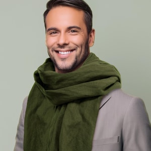 Puede incluir: Un hombre con una chaqueta de traje marrón claro y una bufanda verde oscuro alrededor del cuello. Está sonriendo a la cámara.